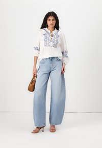 Blouse blanche avec broderie florale bleue, col à volants et cordon de serrage. Associée à un jean large bleu clair et à un sac à main marron.
