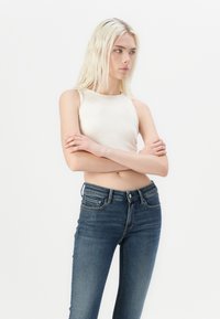 Biely rebrovaný top bez rukávov skombinovaný s tmavomodrými tesnými džínami, ktoré majú klasický dizajn s piatimi vreckami a minimálnymi švovými detailmi.