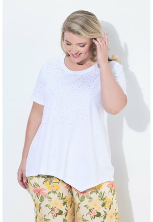 Femme souriante aux cheveux blonds porte un haut blanc à manches courtes avec de petites décorations et un pantalon à motif floral, touchant ses cheveux.
