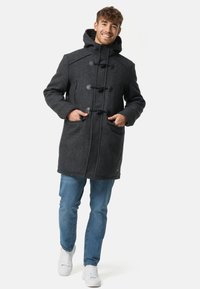 Cappotto grigio scuro in misto lana con cappuccio, chiusure a fibbia, due tasche frontali e vestibilità comoda. Indossato sopra jeans azzurri e sneakers bianche.