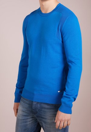 Mężczyzna w jasnoniebieskim swetrze z długim rękawem i wyblakłych niebieskich dżinsach, stojący na tle jasnego, jednolitego tła.