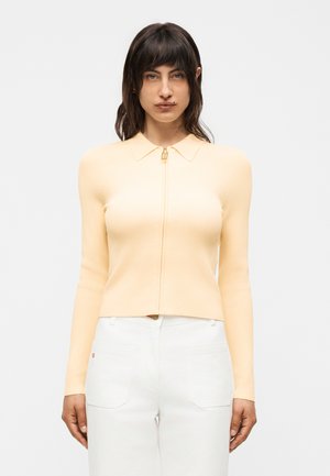 Vrouw met donker haar die een lichtgele zip-up top met lange mouwen en witte broek draagt, staand tegen een effen witte achtergrond.