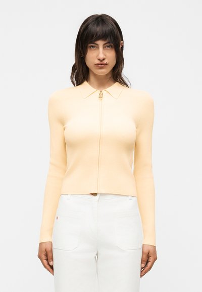Femme aux cheveux foncés portant un haut zippé à manches longues jaune pâle et un pantalon blanc, debout devant un fond blanc uni.