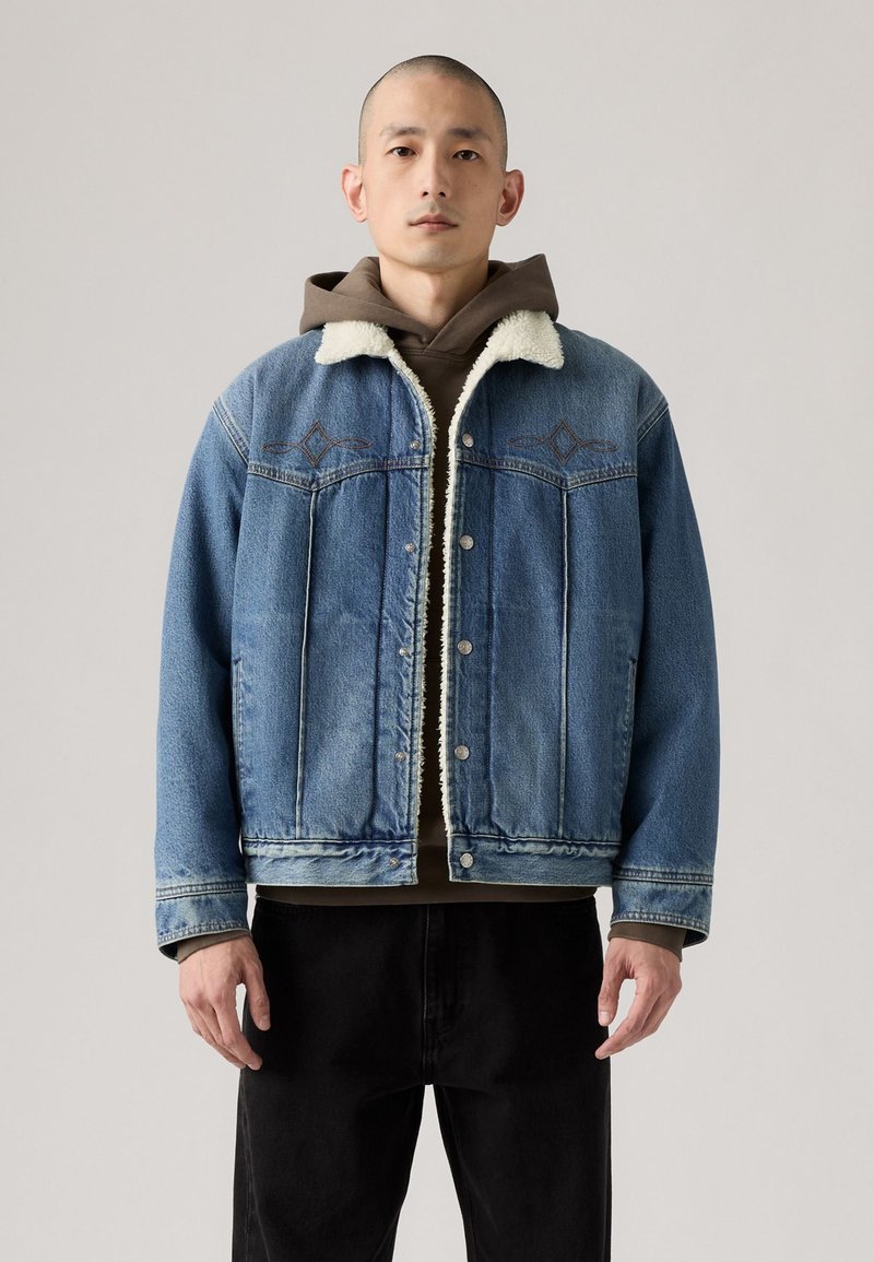 Jasnoniebieska kurtka dżinsowa z kołnierzem wyścielonym Sherpą i ciemnobrązowym wewnętrznym kapturem, charakteryzująca się luźnym krojem i zapięciem na guziki z przodu.