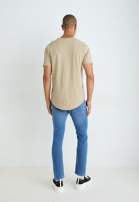 T-shirt en coton beige à manches courtes, ourlet arrondi à l'arrière, associée à un jean bleu et des baskets noires avec des semelles blanches.
