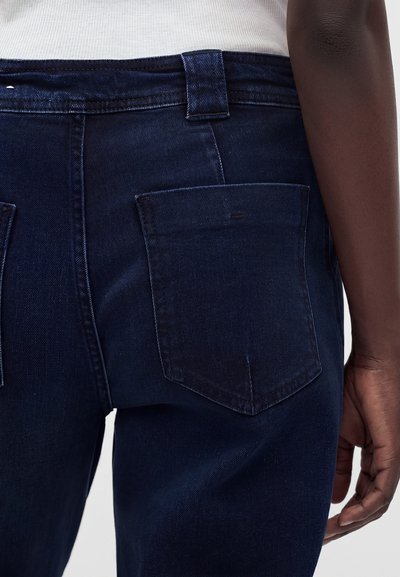 Gros plan de la poche arrière et du côté d'un jean en denim bleu foncé, porté par une personne à la peau foncée et un haut blanc côtelé.