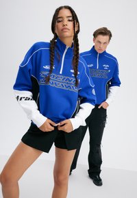 Dwie modelki w dopasowanych niebiesko-białych topach w stylu wyścigowym, jedna w czarnych szortach, druga w podartych czarnych dżinsach, pozują na tle białym.