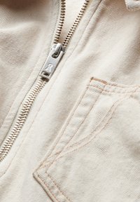 Beige tyg jacka med dragkedja framtill, synliga sömdetaljer och en framlapp. Slät textur med hållbar design.