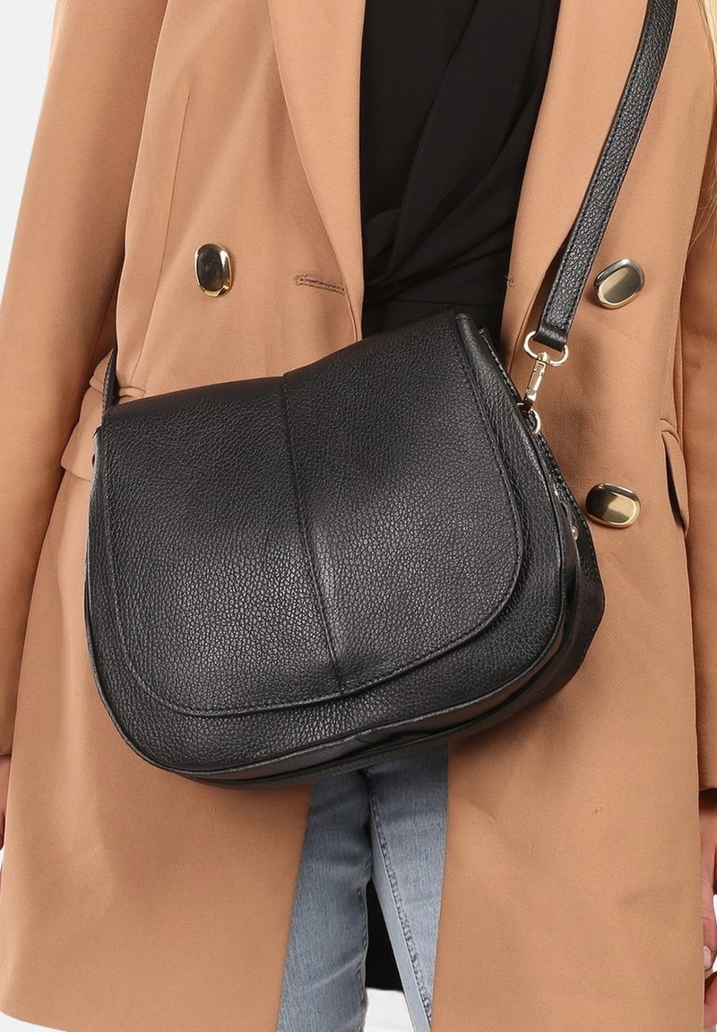 Zwarte leren schoudertas met een gestructureerde afwerking, flap sluiting en verstelbare strap, gedragen over een tan jas en zwart top.