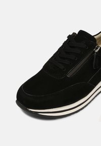 Baskets en daim noir avec lacets noirs, fermeture éclair latérale argentée et semelle rayée blanche et noire sur fond blanc.