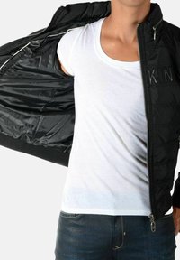 Sort polstret jakke med lynlåslukning foran og ribstrikkede ærmekanter, båret over en hvid T-shirt med rund hals. Denimjeans er synlige forneden.