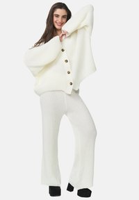 Pull en tricot côtelé blanc avec des manches oversized et une fermeture à boutons, associé à un pantalon évasé en tricot côtelé assorti et des chaussures à plateforme noires.