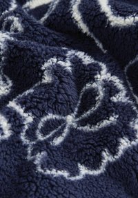 Pătură din fleece de culoare bleumarin cu un model floral alb complicat, având o textură moale și pufoasă și un design ondulat, răsucit.
