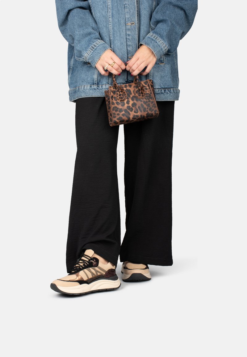 Persona che indossa pantaloni neri a gamba larga, sneakers beige e nere, una giacca di jeans, mentre tiene in mano una piccola borsa a tracolla con stampa leopardata marrone.