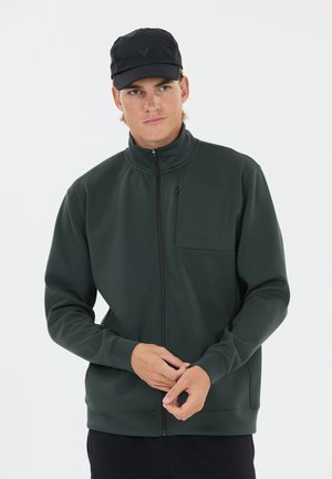 Junger Mann trägt eine dunkelgrüne Jacke mit Reißverschluss und eine schwarze Kappe und passt seine Jackenärmel vor einem schlichten hellen Hintergrund an.