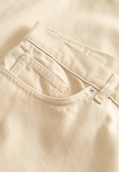 Zbliżenie na jasnobeżowe dżinsy z denimu, pokazujące szlufki na pasek, przednią kieszeń oraz małą kieszonkę na monety z widocznym przeszyciem i srebrnym nitami.