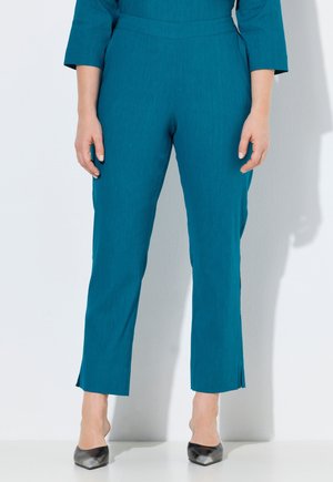 Kvinde iført turkis ankel-lange bukser med sidelommer, kombineret med sorte spidse hæle, stående mod en ensfarvet baggrund.