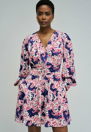 Femme portant une robe à manches longues et col en V, ornée de motifs floraux en spirales roses et bleu marine, debout les mains dans les poches devant un fond uni.