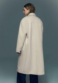 Bež volneni plašč z ohlapnim krojem, širokim ovratnikom in manšetami z gumbi, z enakomerno teksturo in ravnim zadnjim delom.
