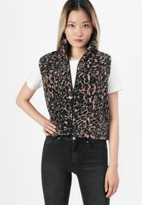 Žena u zatvorenoj, bezrukavnoj prsluku s uzorkom leopard izgleda preko bijele majice i crnih traperica, stoji s jednom rukom na kuku.