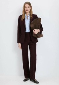 Vinrød skræddersyet dragt med en tætsiddende jakke og bukser med lige ben, kombineret med en lyseblå skjorte og en brun ruskinds taske. Sorte sko.