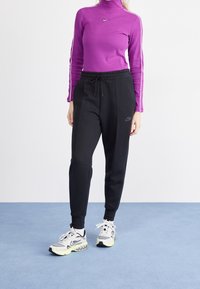 Person som bär en lila långärmad tröja, svarta Nike-joggers och vita Nike-sneakers, står på en ljusblå golv mot en vit bakgrund.