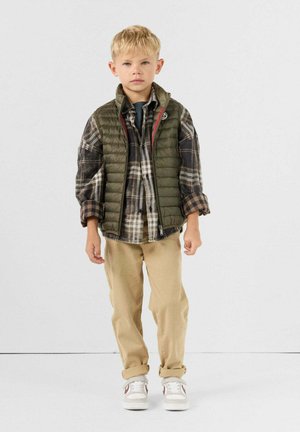 Garçon blond portant une chemise à carreaux, une doudoune verte, un pantalon beige et des baskets blanches, debout devant un fond clair uni.