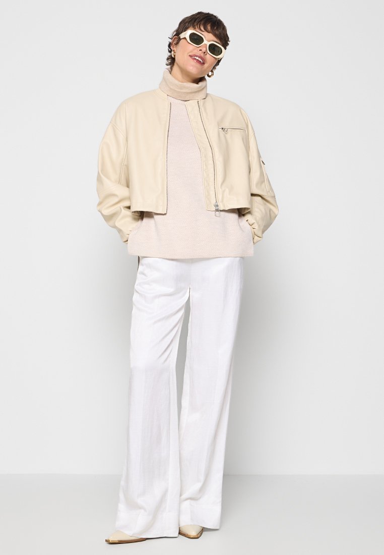 Beige kurz geschnittene Lederjacke mit einem Reißverschluss auf der Vorderseite, kombiniert mit einem hellbeigen Sweater, weißen weit geschnittenen Hosen und beigen spitzen Schuhen.
