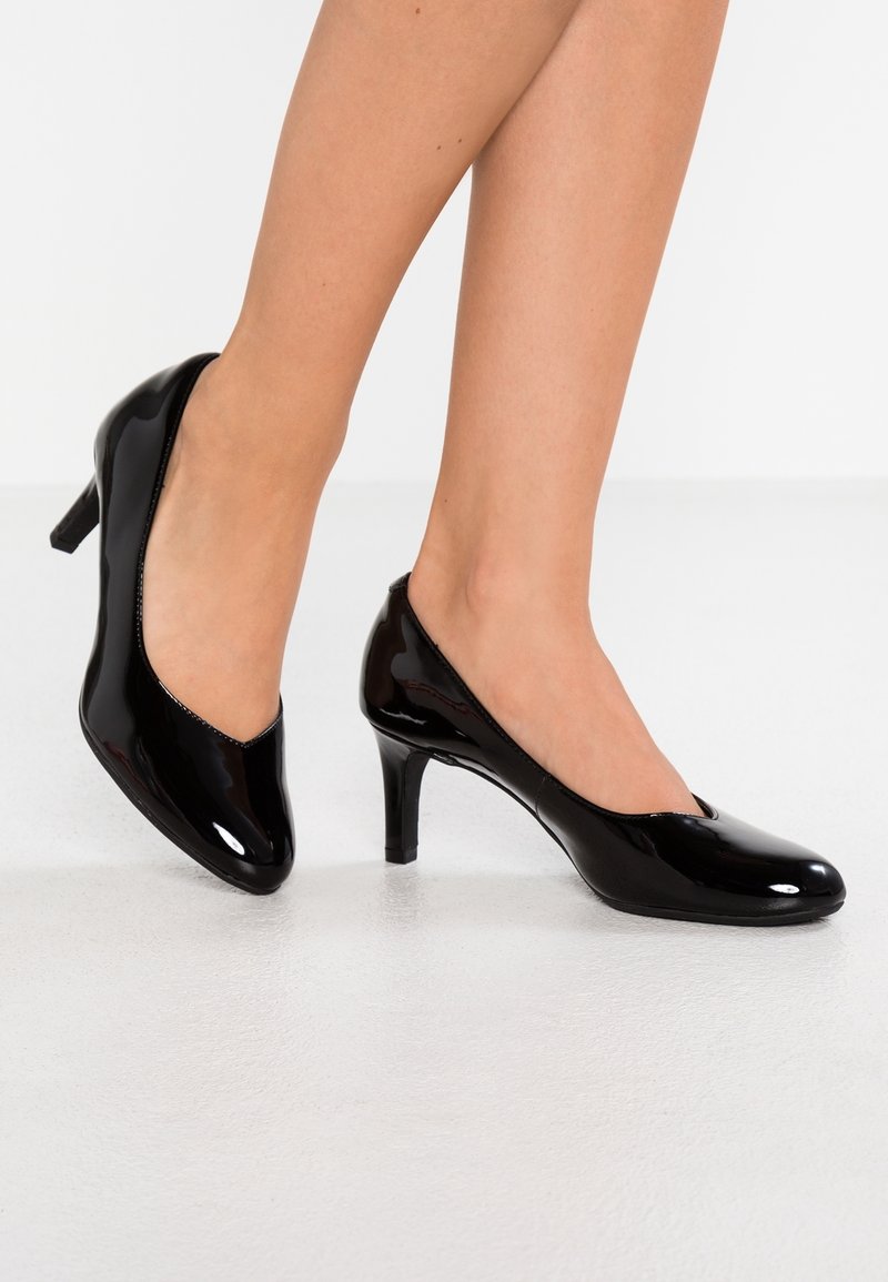 Jambes portant des escarpins à talons moyens en cuir verni noir brillant avec un bout arrondi, sur un sol et un fond blancs.