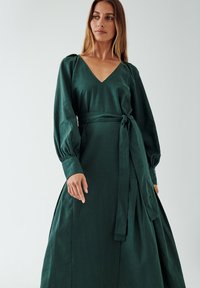 Long green dress made of textured fabric, featuring a V-neck, puffed sleeves, and a tie belt at the waist, with a flowing skirt design.