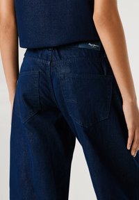 Tmavomodré džíny z denimu s vysokým pásom a dizajnom s piatimi vreckami. Majú jemný lesk a kontrastné švy. Na zadnej strane je viditeľná logo nálepka.