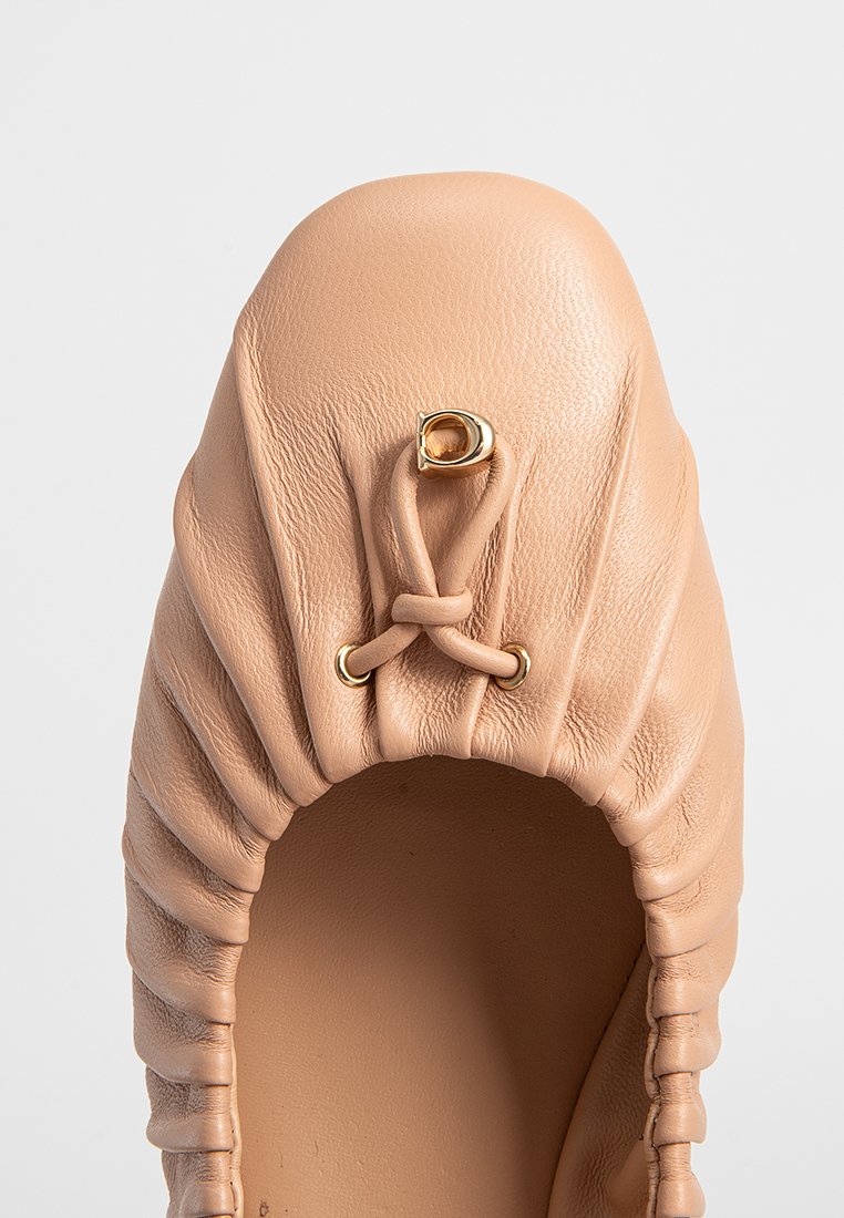 Nude leren balletschoen met geplooide textuur, ronde neus en een gouden hardwareaccent. Het interieur is glad en onversierd.