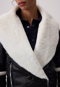 Sieviete, kura valkā melnu ādas jaku ar lielu baltu vilnas apkakli virs tumši zilas pogas krekla, kas ir apgriezts zem deguna.