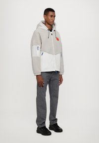 Giacca con cappuccio grigia e bianca in tessuto felpato, con tasche con zip e dettagli blu; abbinata a jeans grigio scuro e sneaker nere.