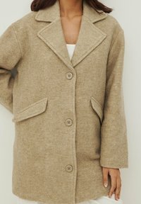 Béžový vlněný kabát s klopovým límcem, třemi velkými knoflíky a dvěma předními kapsami. Hladká textura a volný střih.