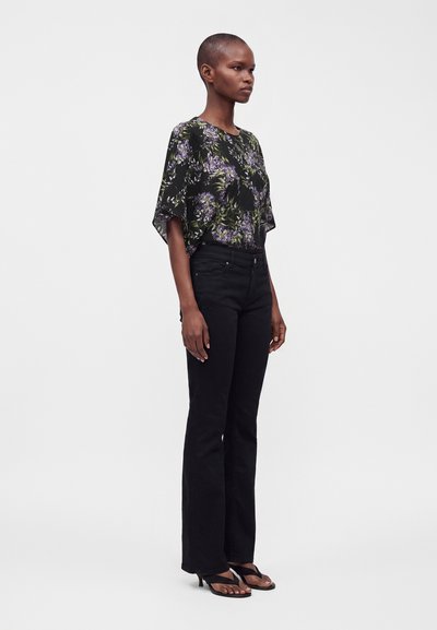 Femme debout portant un chemisier noir à motifs floraux avec des fleurs violettes, un pantalon noir taille haute et des sandales à talons noires sur fond blanc.