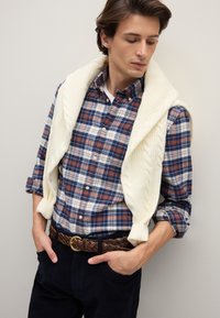 Herrenausstattung mit einem blau-roten karierten Hemd, cremefarbenem, strickgestricktem Pullover, der über den Schultern drapiert ist, braunem geflochtenem Gürtel und dunkler Hose.