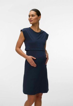 Femme enceinte portant une robe sans manches bleu marine, les mains reposant doucement sur son ventre, regardant sur le côté sur un fond blanc uni.