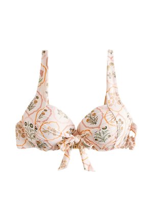 Kobieta o długich blond włosach, ubrana w bikini w kwiatowy wzór, stoi na zewnątrz obok drewnianego stołu z owocami i szklankami.