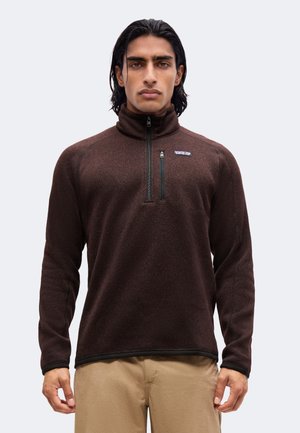 Uomo con capelli scuri che indossa un pullover in pile marrone con zip al quarto e pantaloni beige, in piedi davanti a uno sfondo semplice.