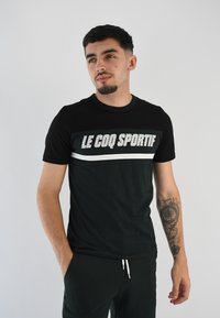 Camiseta negra con un logo gris de "LE COQ SPORTIF" en el pecho, de mangas cortas y cuello redondo. La tela parece suave y lisa.