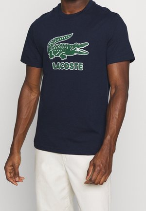 Homme portant un t-shirt bleu marine à manches courtes avec le logo crocodile vert de Lacoste et un texte blanc, assorti à un pantalon blanc cassé.