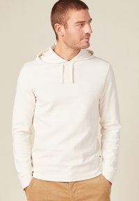 Helle beige Kapuzenshirt mit langen Ärmeln, strukturiertem Stoff, Fronttasche und Kordelzugkapuze. Kombiniert mit khakifarbenen Hosen.