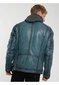 Läderjacka i en mörk teal-färg med strukturerad yta, kontrasterande sömmar och en grå huva som foder. Utrustad med sömdekor.