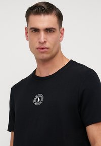 Czarna koszulka wykonana z bawełny, z okrągłym dekoltem, krótkimi rękawami oraz okrągłym haftowanym logo na piersiach. Gładka tekstura.