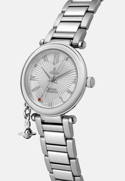 Montre en acier inoxydable argenté avec un cadran rond en forme de cœur et des chiffres romains. Comprend un détail rouge et un pendentif sur le côté.