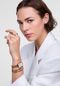Montre en or avec un cadran carré et un bracelet en maille au poignet, accompagnée de trois bracelets en or fins avec des accents circulaires. Fond de blazer blanc.