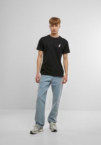 Svart bomull t-skjorte med korte ermer, med en liten fargerik grafikk på venstre bryst. Sammen med lyseblå jeans og sneakers.