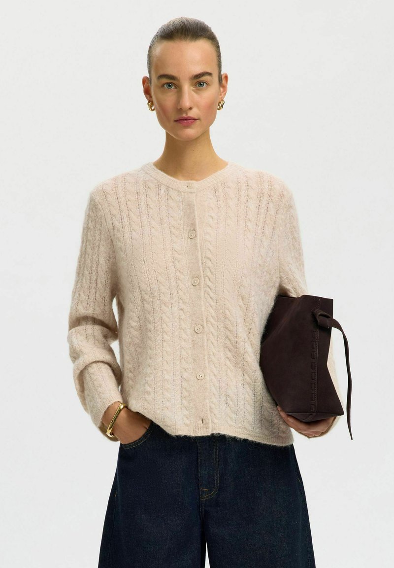Femme aux cheveux attachés en chignon porte un cardigan beige en maille torsadée et un jean foncé, tenant un sac à main en daim marron foncé avec une lanière dans une main.