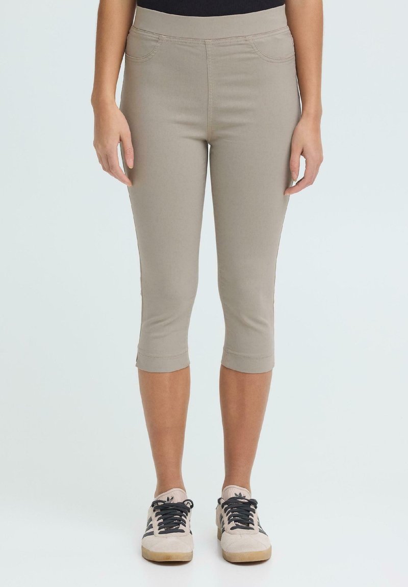 Γυναίκα που φοράει μπεζ παντελόνι cropped και ανοιχτόχρωμα αθλητικά παπούτσια με μαύρες κορδόνια, στέκεται μπροστά σε απλό φόντο.